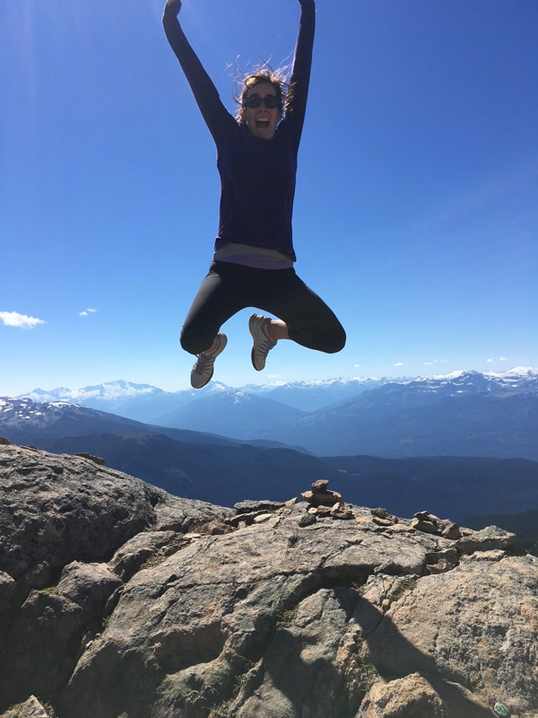 Person jumps in the air on a rocky mountain overlook with snow-capped peaks and blue sky in the background.