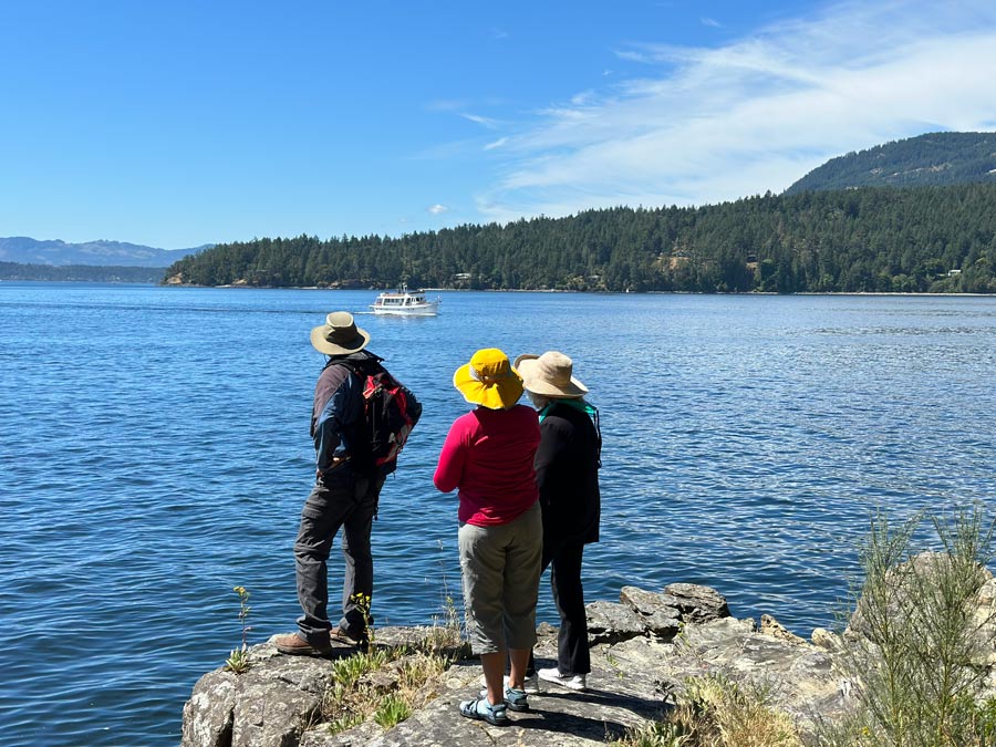 Three hikers stand along a rocky shoreline overlooking a calm lake with forested hills and a boat in the distance.