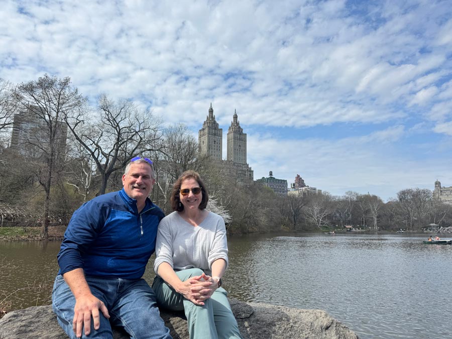Two people sit together on a rock overlooking a lake in Central Park with the New York City skyline visible in the background.
