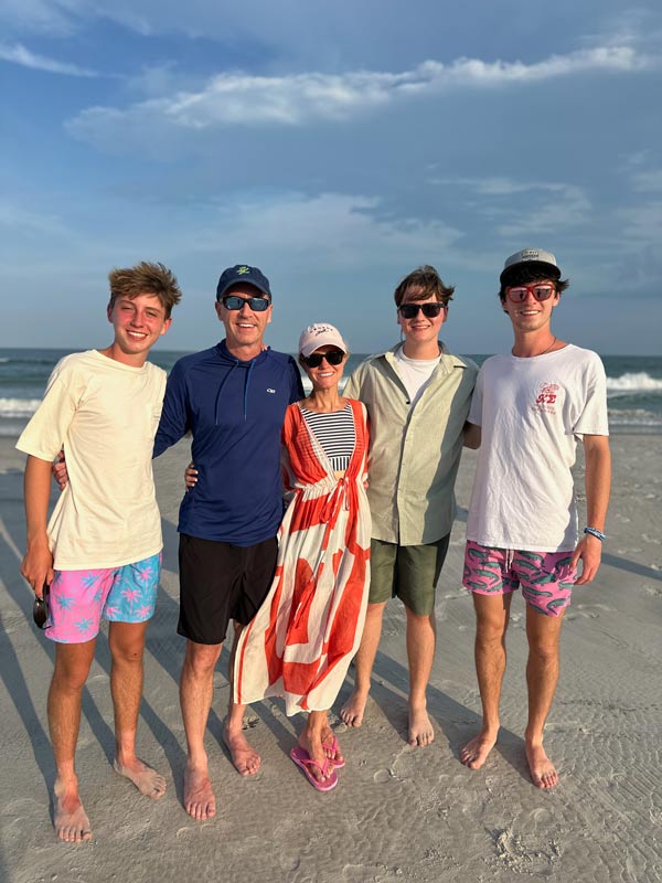 Five people stand together barefoot on a sandy beach at sunset, smiling toward the camera with ocean waves and a cloudy sky in the background.