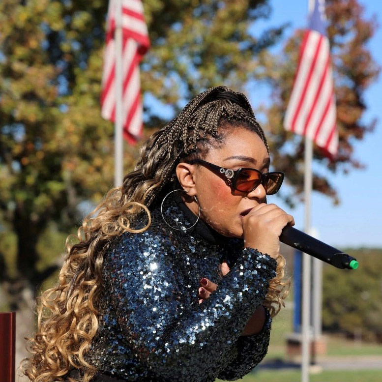 Performer wearing a sequined jacket and sunglasses sings into a microphone outdoors with American flags displayed in the background.