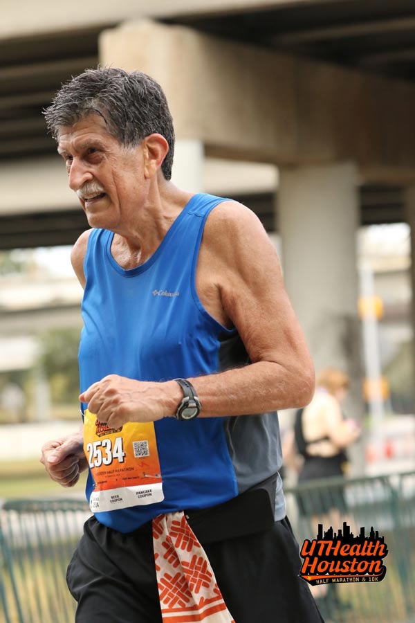 A runner wearing a blue tank top and race bib participates in the UTHealth Houston Half Marathon & 10K with an overpass visible in the background.