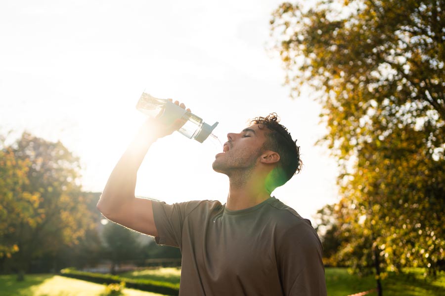 A man in a gray shirt drinks water in a field with trees as the sun shines down on him