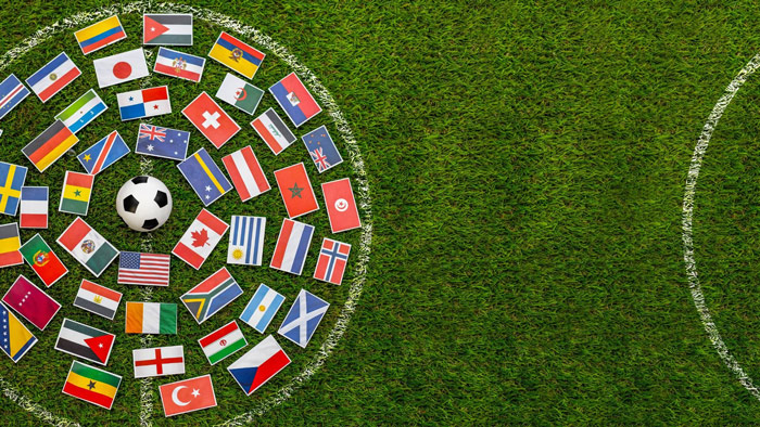 Soccer ball surrounded by miniature international flags arranged in a circle on a grass field, creating a global soccer theme with open space on the right side of the image.