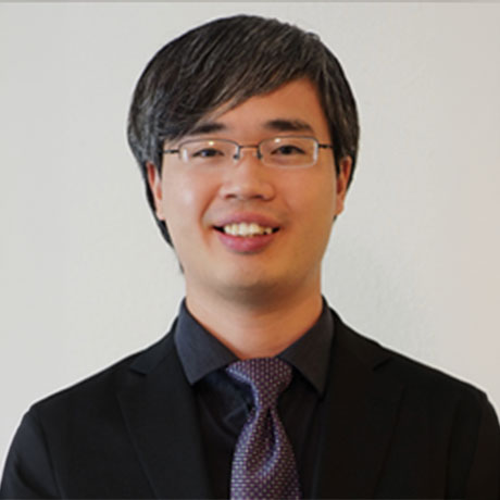 Person wearing glasses, a dark suit, and a patterned tie, smiling at the camera.