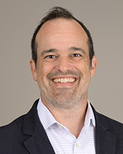 Dr. Matthew Baker poses for a professional headshot in black suit and white button up in front of a gray background
