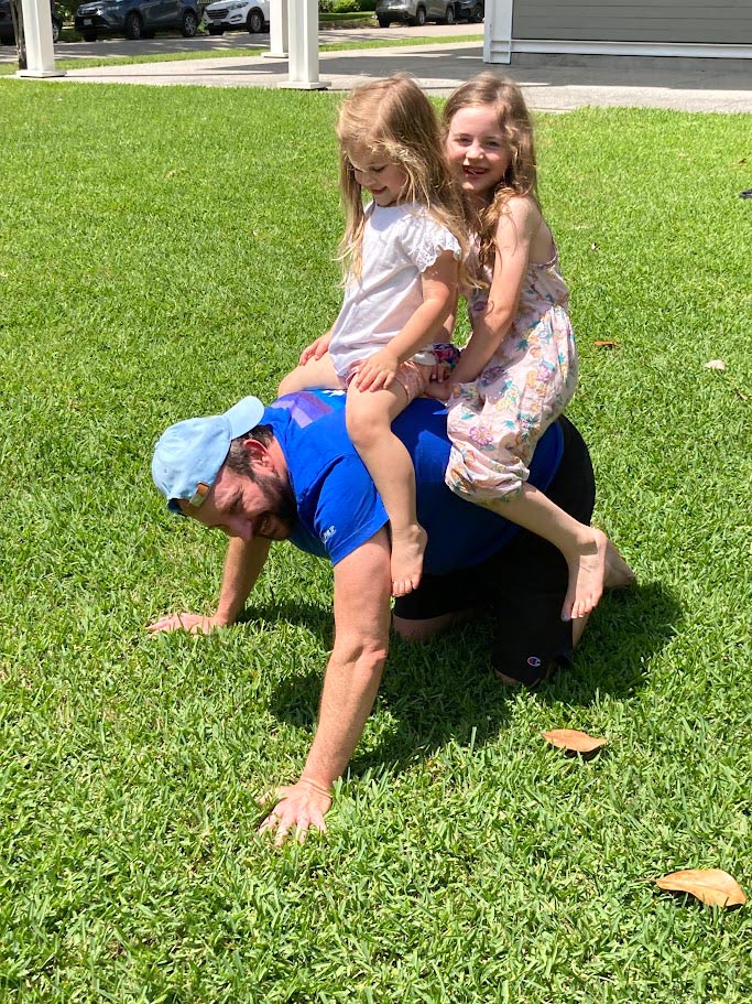 Two young girls laugh as they ride on the back of an adult crawling across a grassy lawn during outdoor play in a neighborhood yard.
