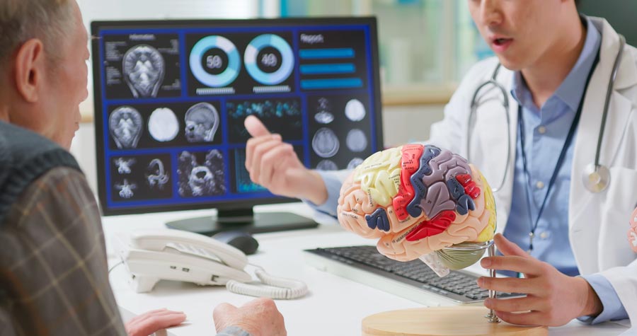 A physician wearing a white coat explains a color-coded brain model to an older patient during a consultation. Brain scans and diagnostic images are displayed on a computer monitor in the background as the physician gestures while discussing the patient's condition.