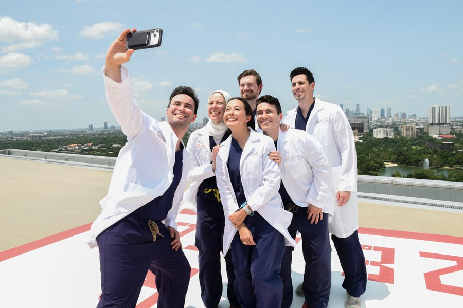Six chief residents from the Department of Orthopedic Surgery at McGovern Medical School at UTHealth Houston gather on the rooftop helipad at Memorial Hermann Hospital as one resident takes a group selfie, with the Houston skyline stretching behind them on a sunny day.