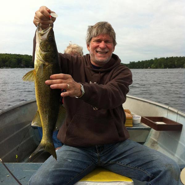 Dr. Ronald Rapini smiles while sitting in a fishing boat on a lake in Canada, holding up a large walleye he caught. Trees line the distant shoreline under a lightly clouded sky.