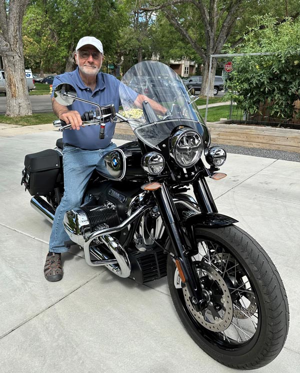A man sits on a black BMW R 18 motorcycle in a residential driveway, smiling at the camera while wearing a blue polo shirt, jeans, and a white cap.