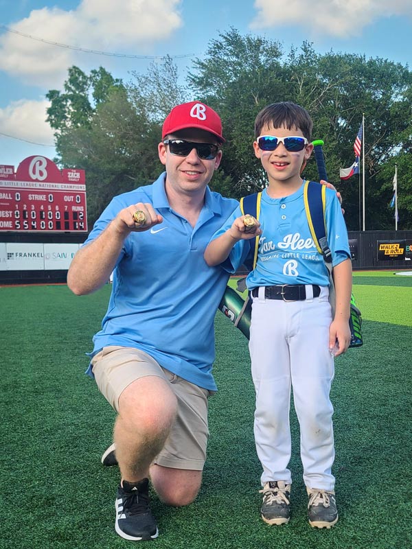 An adult and a young baseball player wearing Bellaire Little League uniforms pose on the field while proudly displaying championship rings after a game.