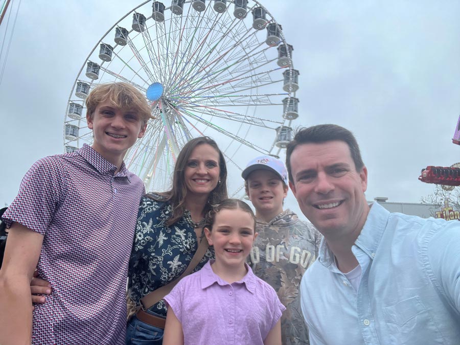Dr. Ryan Hall and his family smile for a selfie at the Houston Rodeo with a Ferris wheel rising behind them. The group stands together in front of the carnival attractions on an overcast evening.