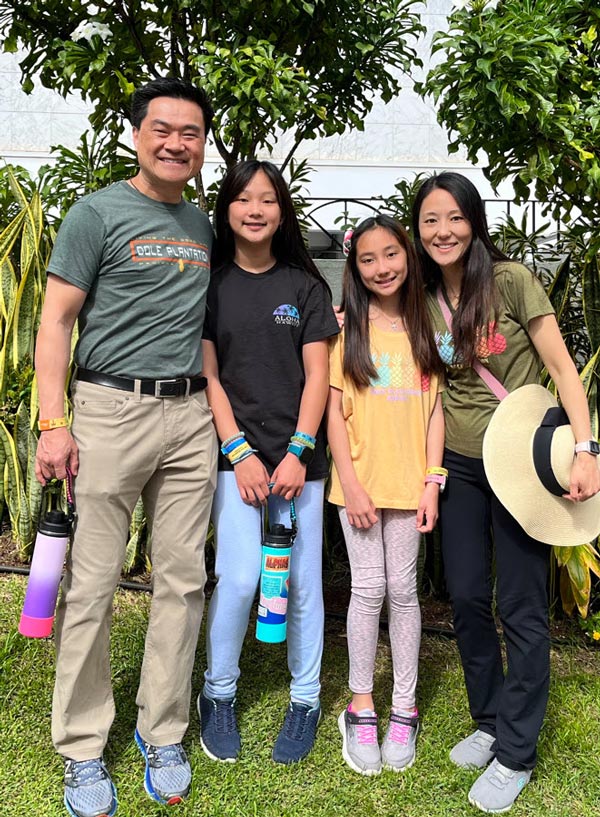 Dr. Peggy Hsieh poses with her husband and two daughters in a lush outdoor garden during a family trip to Taiwan. The family smiles together beneath flowering trees.