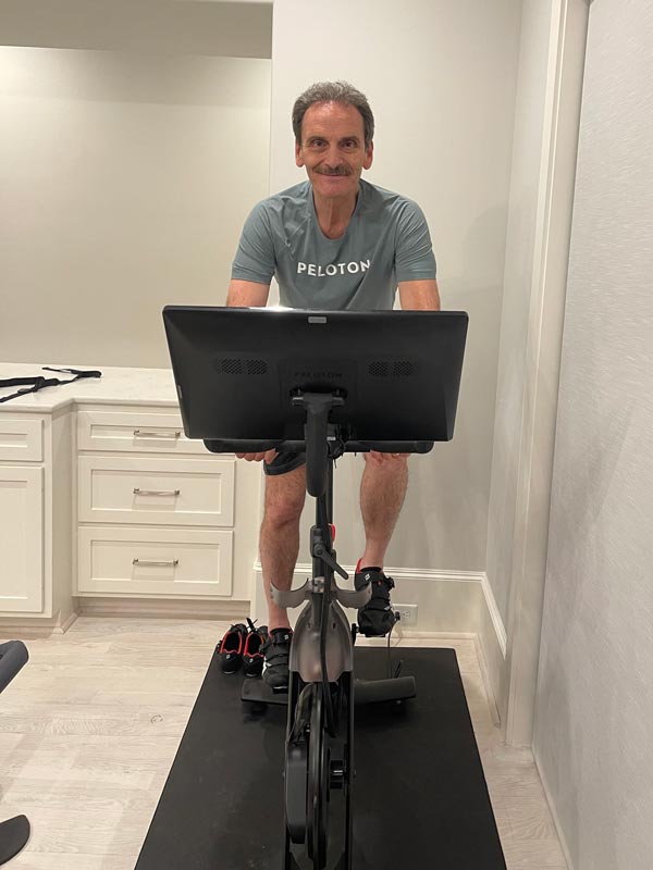 Dr. Jacques Morcos smiles while riding a Peloton exercise bike in a bright home workout space, wearing a Peloton T-shirt and cycling shoes.