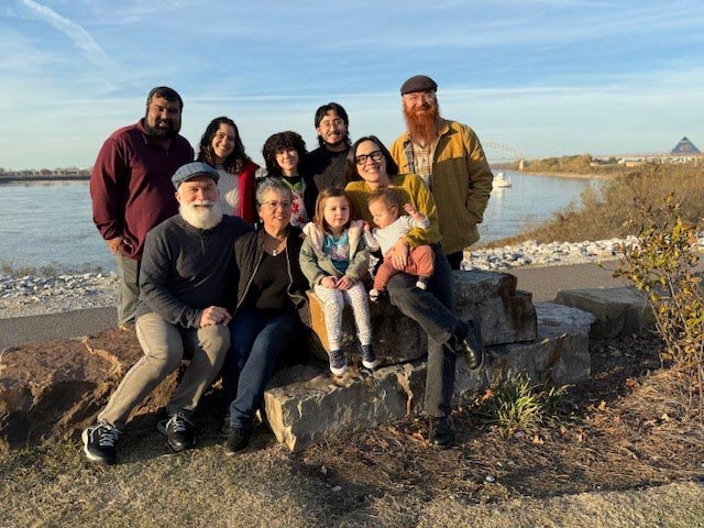 A multigenerational family poses together on large rocks overlooking the Mississippi River at Tom Lee Park in Memphis, Tennessee, with the Hernando de Soto Bridge visible in the background during golden hour.