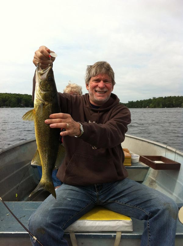 Dr. Ronald Rapini smiles while sitting in a fishing boat on a lake in Canada, holding up a large walleye he caught. Trees line the distant shoreline under a lightly clouded sky.