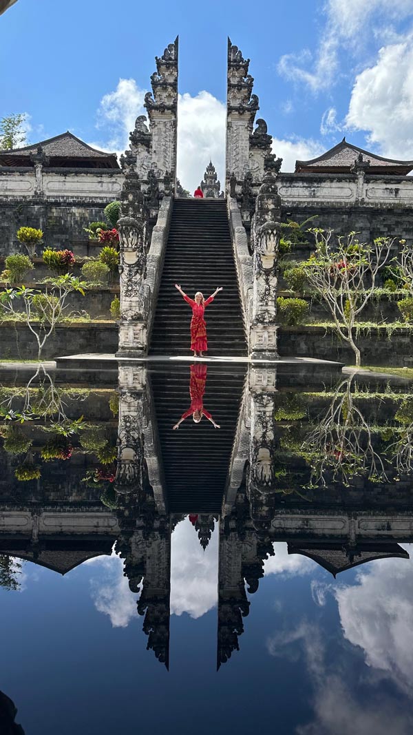 A woman in a red dress stands with her arms outstretched at the base of the grand staircase of a Balinese temple, reflected in a still pool beneath a bright blue sky.