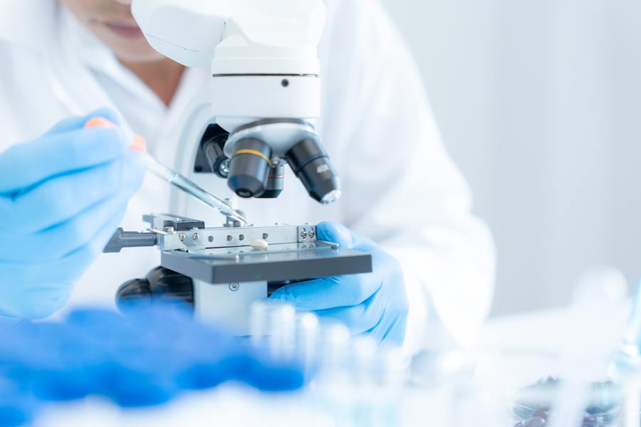 Close-up of a researcher wearing blue gloves using a microscope while carefully manipulating a specimen on a glass slide with laboratory tools. Blurred laboratory equipment in the foreground emphasizes the biomedical research setting.