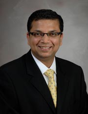 Dr. Nirav Thosani poses for a professional headshot in a black suit with yellow tie in front of a gray brackground.