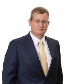 Brian Dean poses for a professional headshot in a black suit, white shirt, and yellow tie