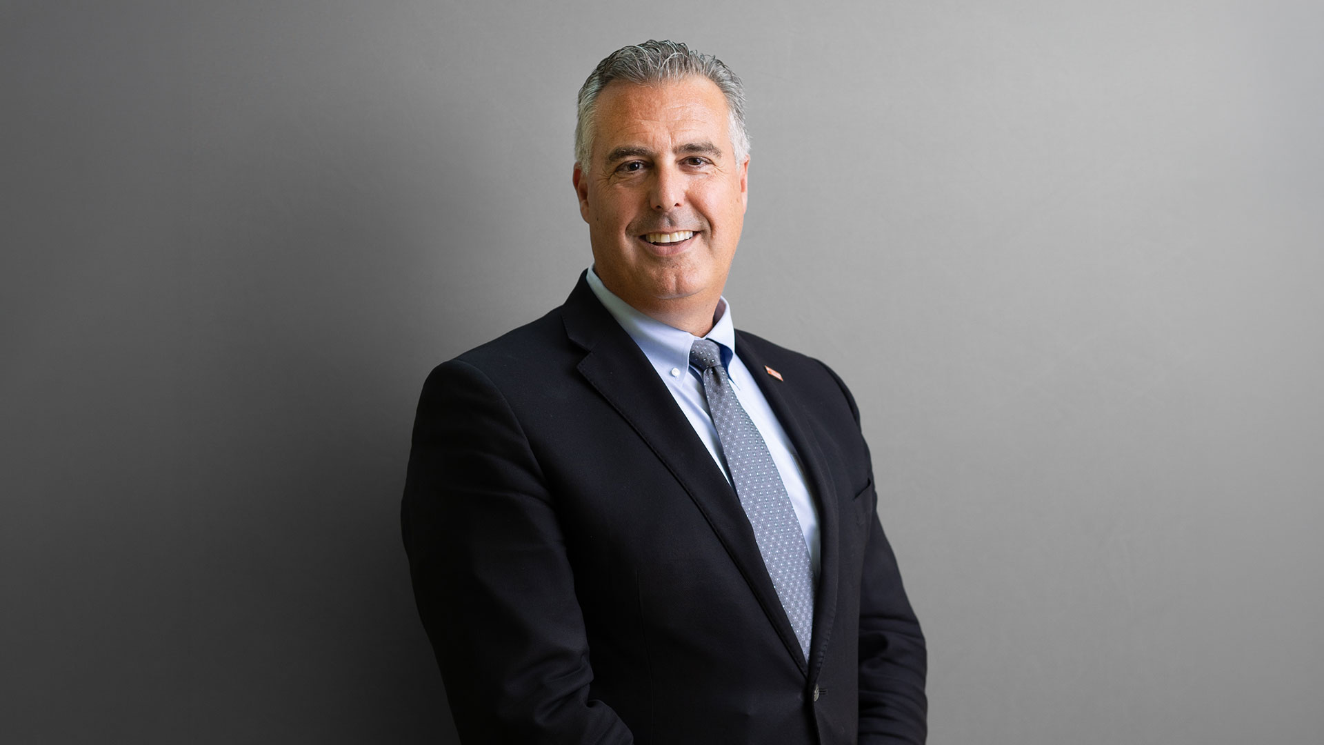 Dr. Kevin Morano poses for a professional headshot in a black suit, blue shirt, and blue tie