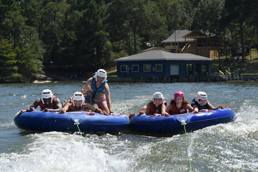 Five medical students wearing helmets and life jackets ride two inflatable towable tubes across a lake during the Henry Strobel Retreat, smiling as a boat pulls them over the water with lakeside cabins and trees in the background.