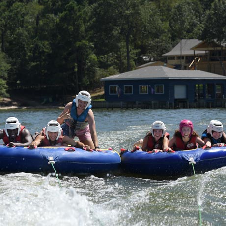 Five medical students wearing helmets and life jackets ride two inflatable towable tubes across a lake during the Henry Strobel Retreat, smiling as a boat pulls them over the water with lakeside cabins and trees in the background.