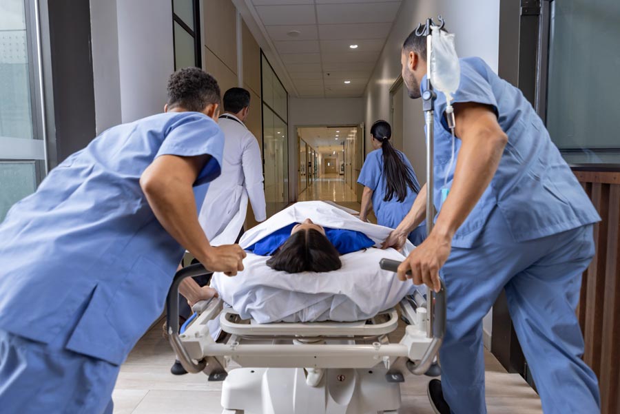 Medical team members in blue scrubs rush a patient on a hospital bed down a hallway while an IV bag hangs beside the bed.