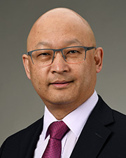 Dr. Samden Lhatoo poses for a professional headshot in a black suit with white shirt and maroon tie