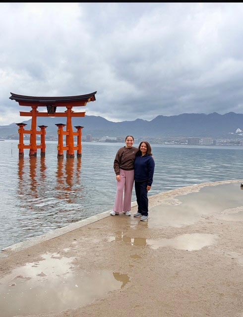 Bela Patel, MD, and a family member stand along the shoreline in Japan with a large red torii gate rising from the water behind them.