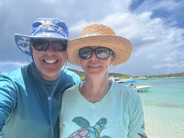 Angela Smith, CPA, and her husband, Andy, smile for a selfie while wearing sun hats and sunglasses beside the turquoise waters of Culebrita, Puerto Rico.