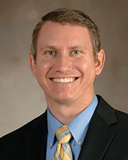 Dr. Devin McBride poses for a professional headshot with a black suit, blue shirt, and yellow and blue tie
