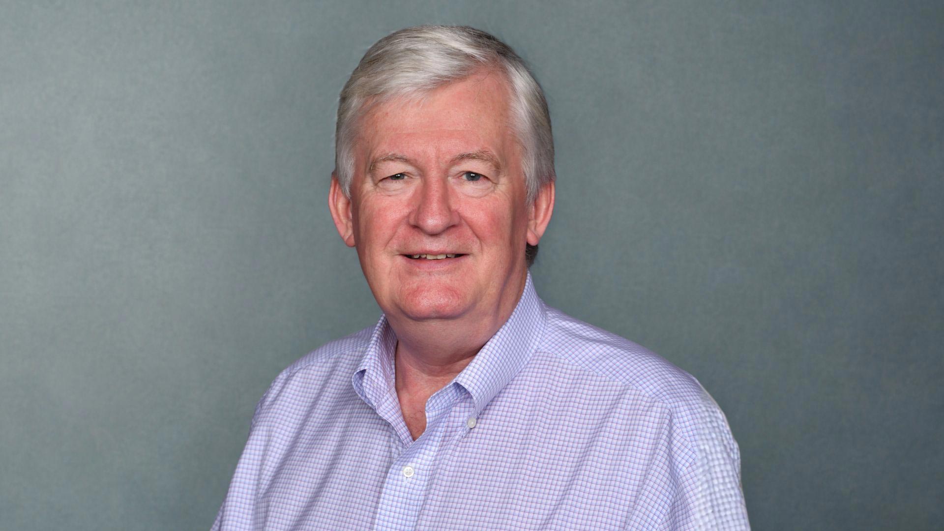 Dr. Peter Doris poses for a professional headshot in a purple button up shirt in front of a gray background