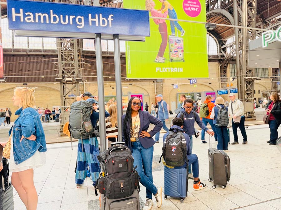 A woman stands smiling with rolling luggage beneath a Hamburg Hauptbahnhof station sign in Germany as travelers move through the busy train station.