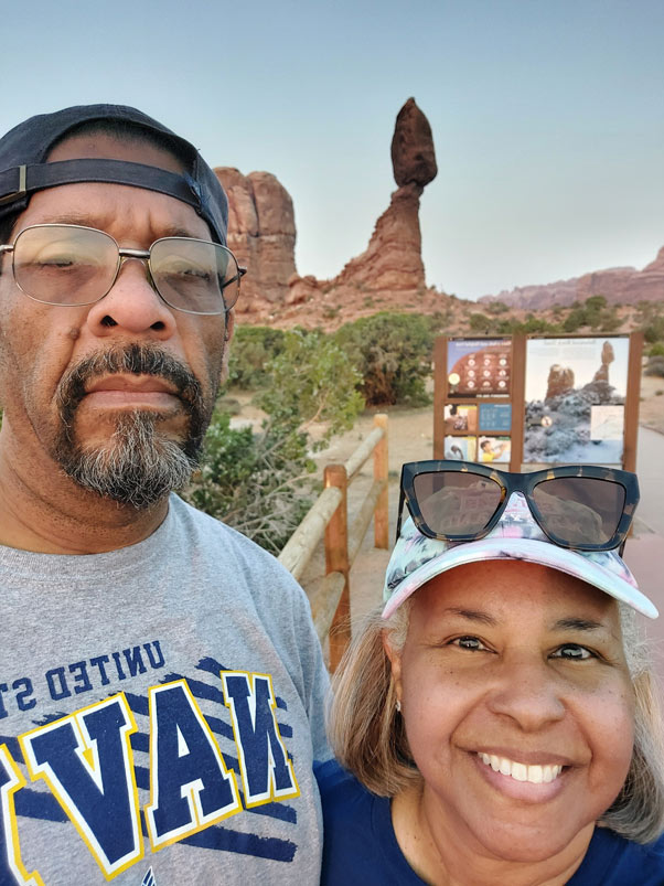 A couple takes a selfie at Arches National Park in Moab, Utah, with the distinctive Balanced Rock formation and red rock landscape visible behind them.