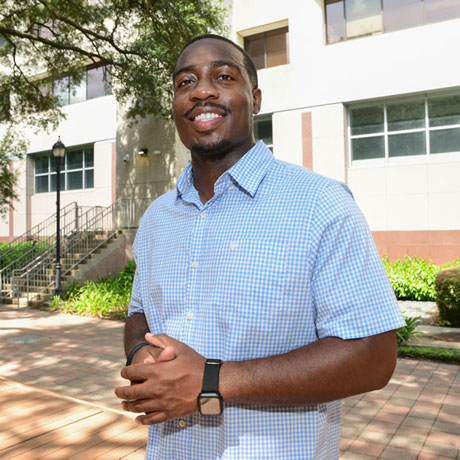 McGovern Medical School student Jeremiah Ageh poses for a photo in front of the TMC Library in a blue button up shirt