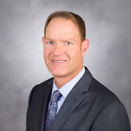 Duncan Campbell poses for a professional headshot in a gray suit with blue button up shirt and blue tie