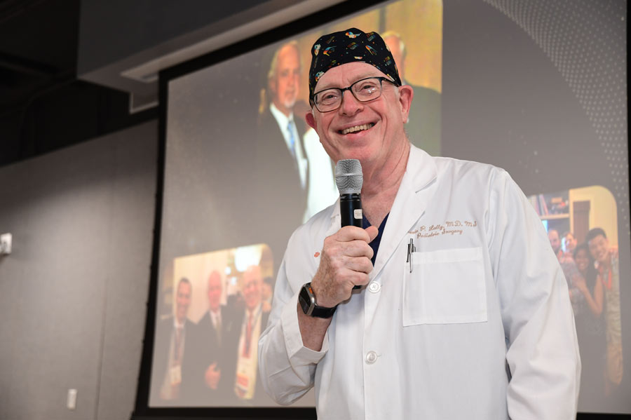 A physician in a white coat and surgical cap speaks into a microphone during Richard Andrassy, MD’s retirement celebration.