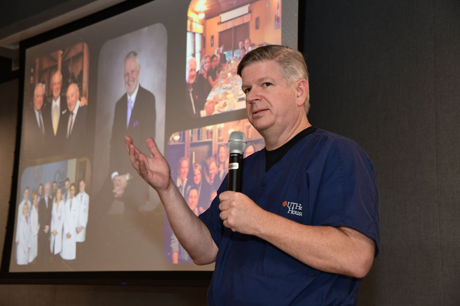 A physician in surgical scrubs speaks into a microphone during Richard Andrassy, MD’s retirement celebration, with photos from Andrassy’s career displayed behind him.