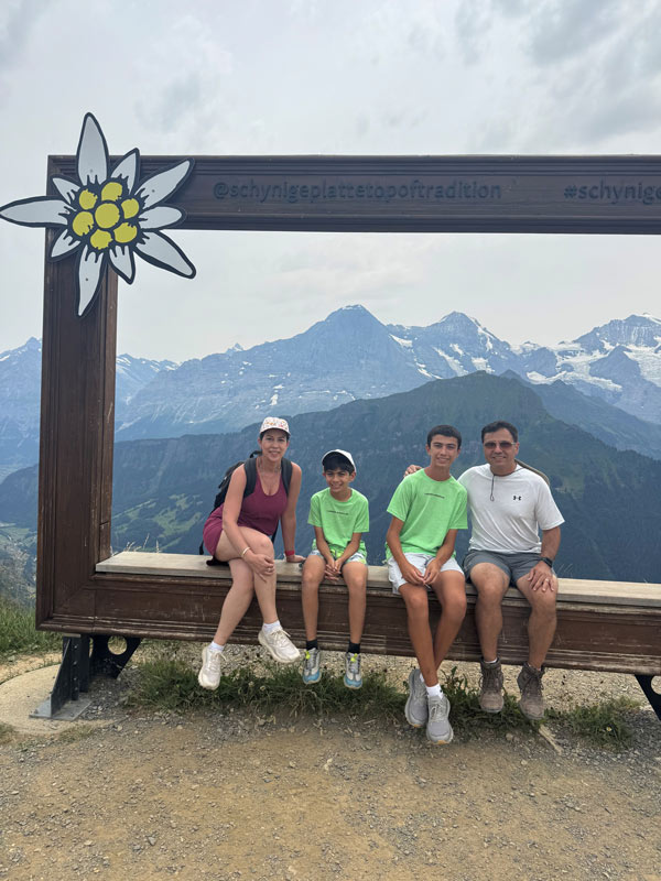 A family of four sits inside a large wooden picture frame overlooking the Swiss Alps, with green mountain slopes and snow-covered peaks in the background.