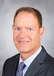 Duncan Campbell poses for a professional headshot in a gray suit with blue button up shirt and blue tie