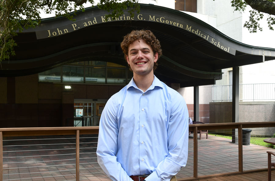 Connor Dechiro poses for a photo in a blue button up shirt in front of an awning that reads McGovern Medical School