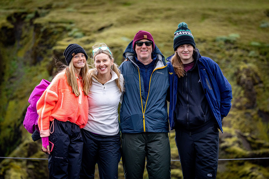 A family of four poses together outdoors in Iceland, bundled in jackets and hats against a green, rugged landscape.