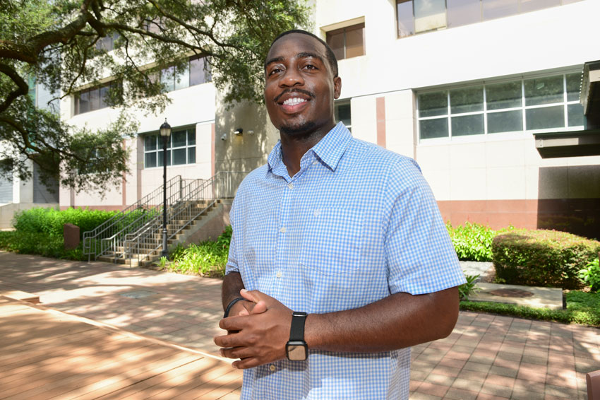 McGovern Medical School student Jeremiah Ageh poses for a photo in front of the TMC Library in a blue button up shirt