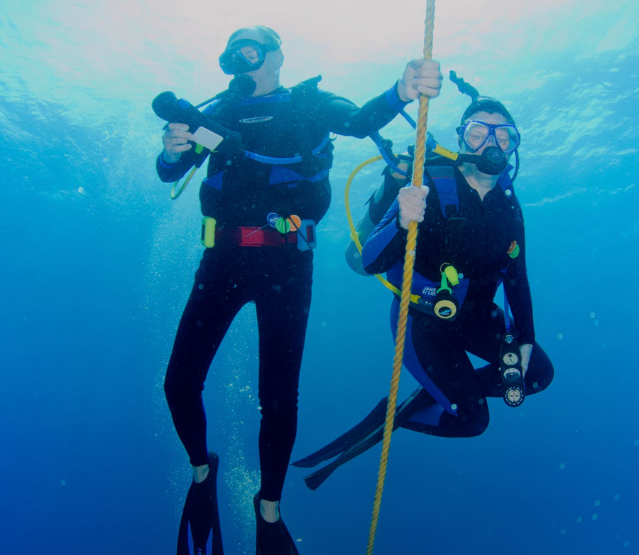 Two scuba divers in diving gear hold onto a rope while underwater in the clear blue waters of Grand Cayman.
