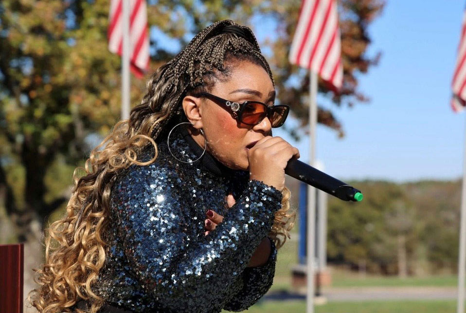Performer wearing a sequined jacket and sunglasses sings into a microphone outdoors with American flags displayed in the background.