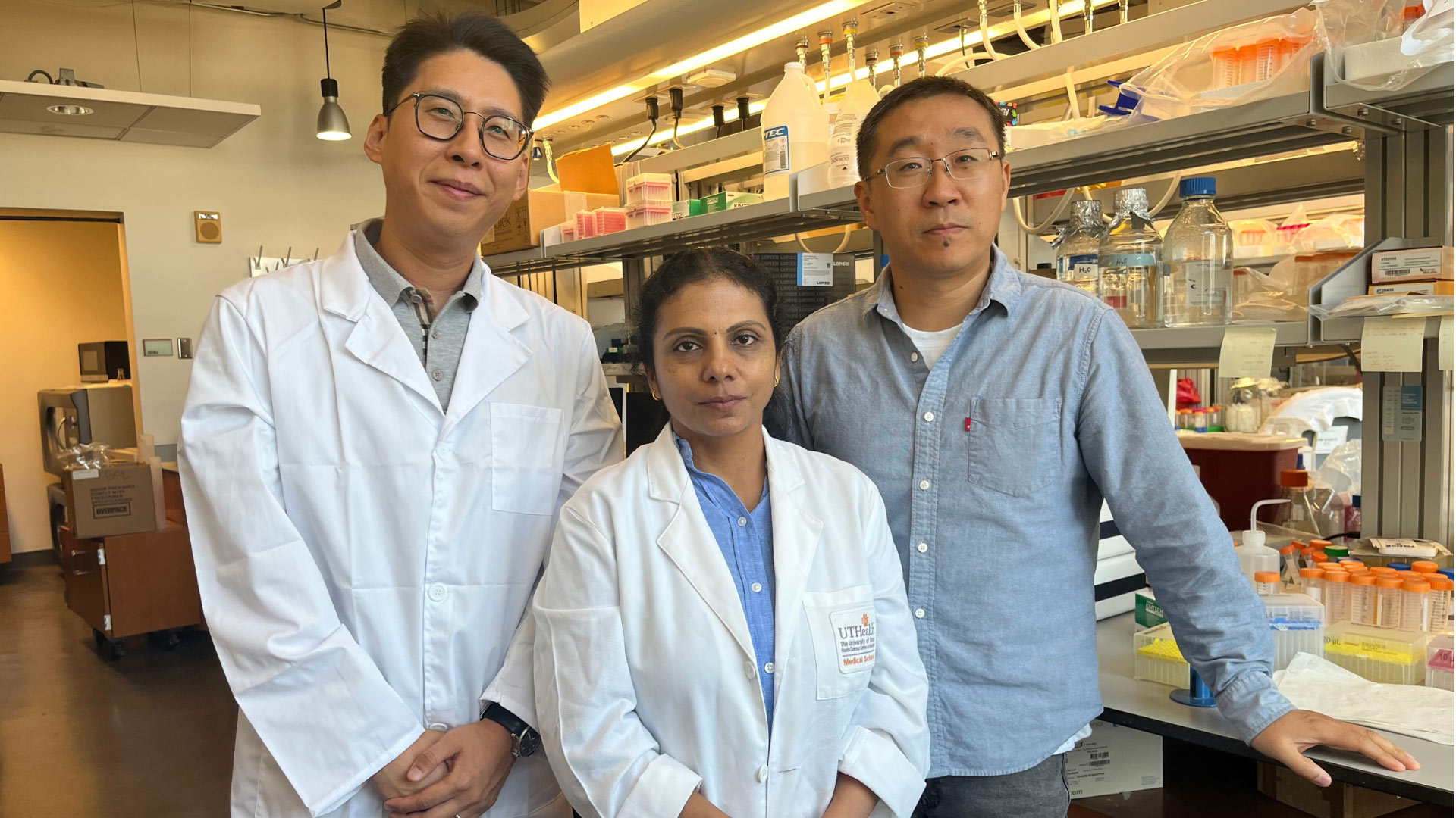 Three researchers pose together in a laboratory, with two wearing white lab coats and laboratory equipment and supplies visible in the background.