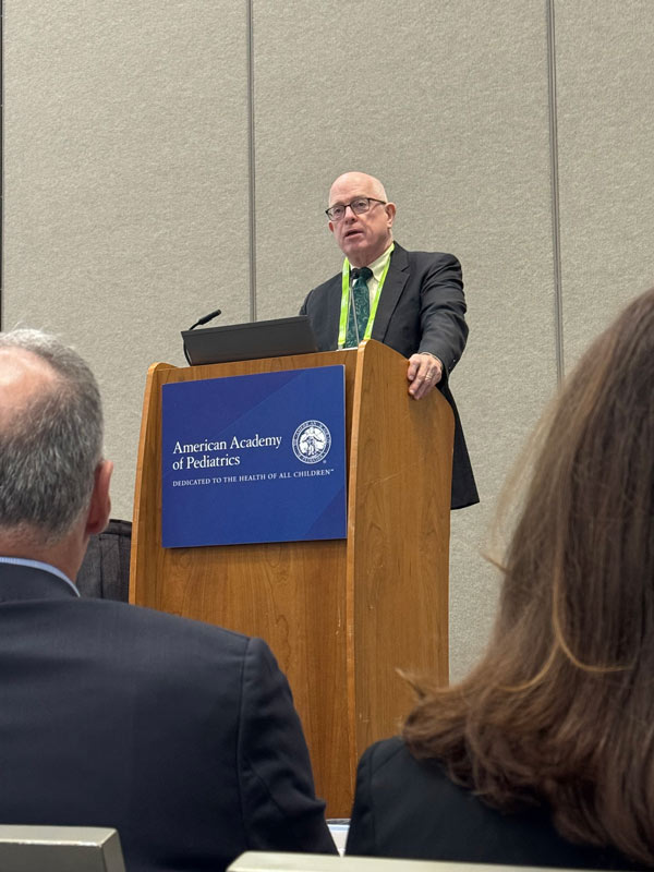 Kevin Lally, MD, MS, speaks from a wooden podium displaying the American Academy of Pediatrics logo during a conference presentation.